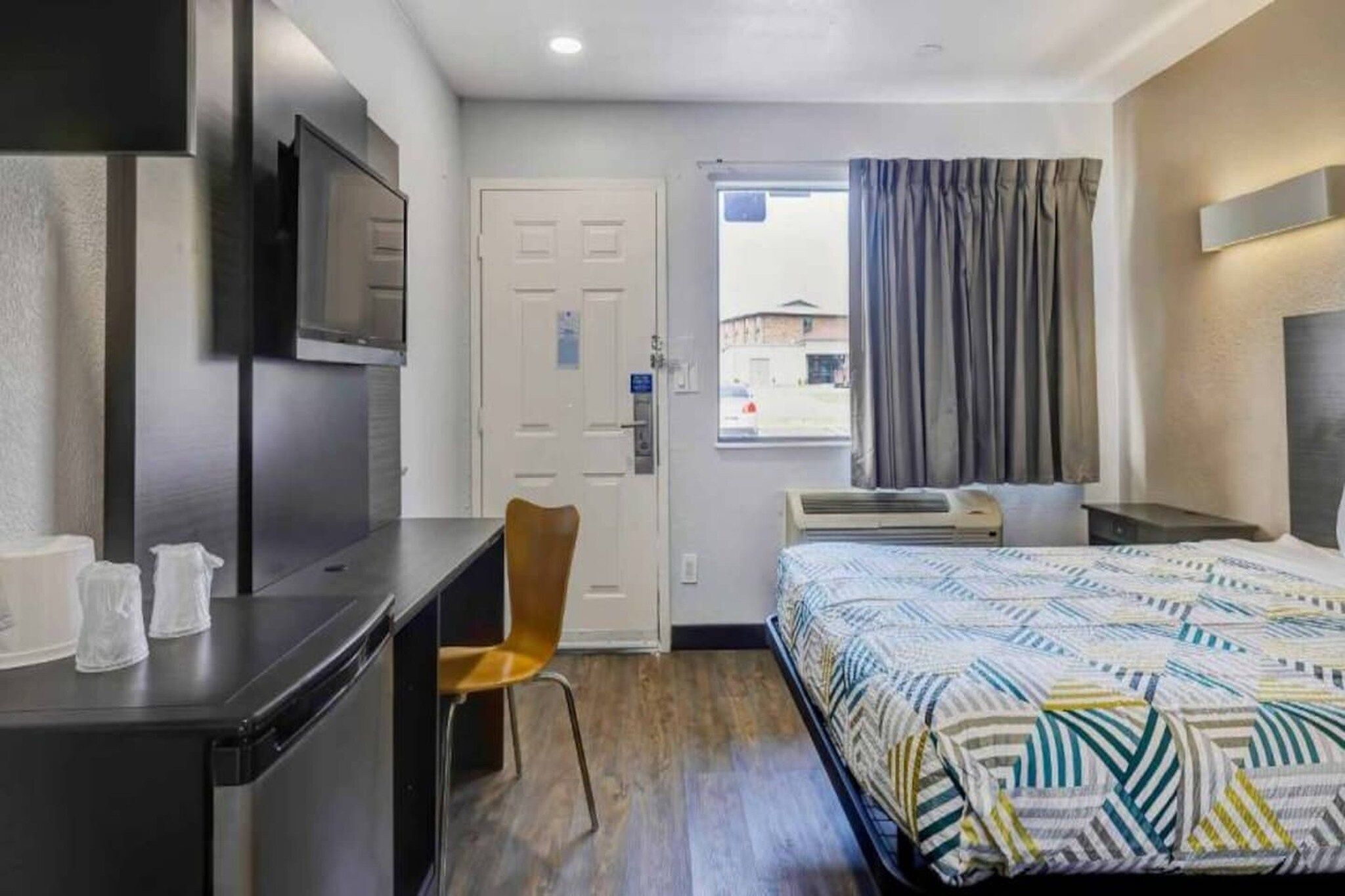 A hotel room with a bed, a desk, a chair, a TV, a window with curtains, and an air conditioning unit.