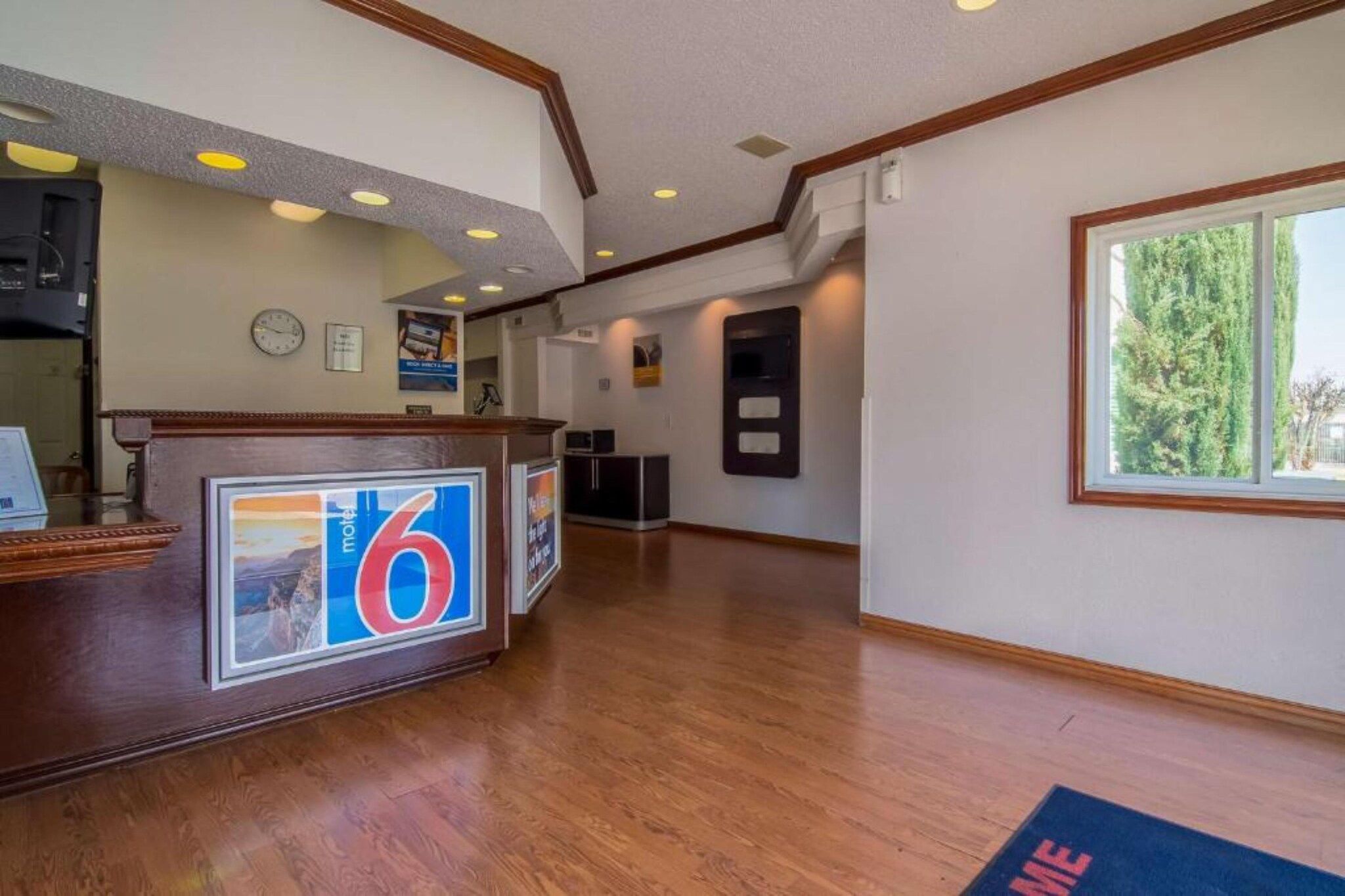 A hotel reception area with a wooden reception desk, a clock, and a window with a view of trees.