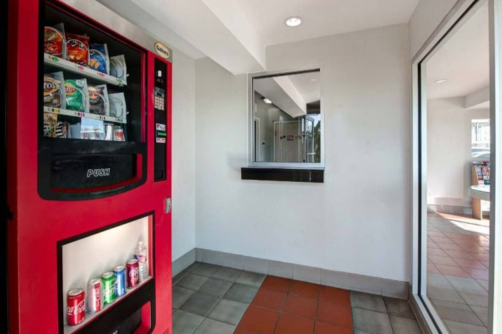 A hallway with a vending machine stocked with snacks and drinks, a mirror on the wall, and a tiled floor.