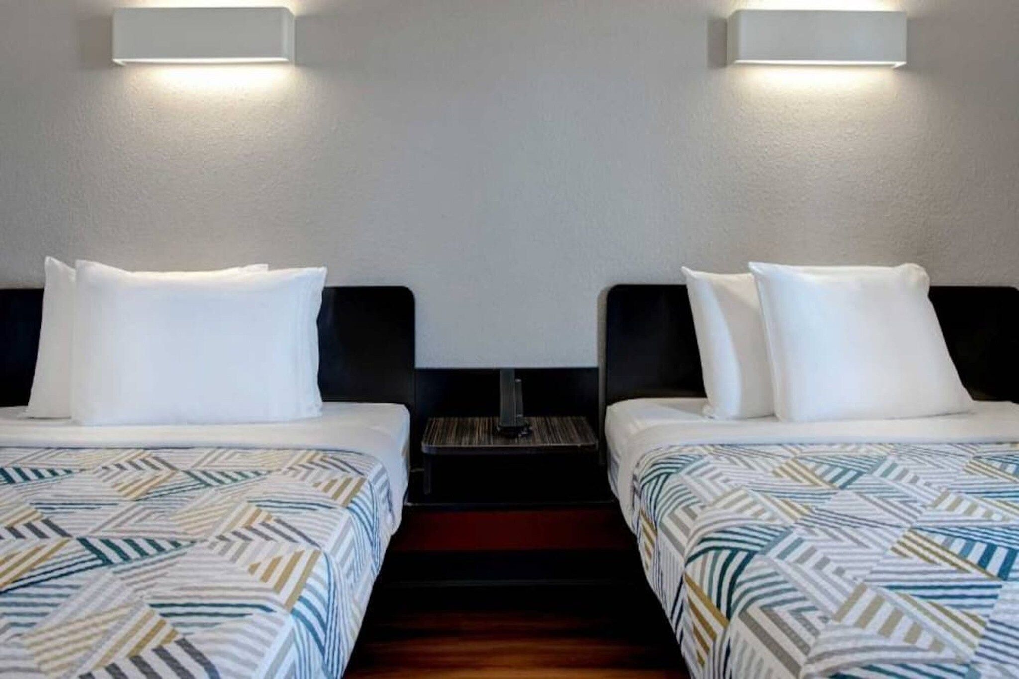 Een hotelkamer met twee bedden, elk voorzien van een bedsprei met een patroon en witte kussens, en een nachtkastje ertussen.