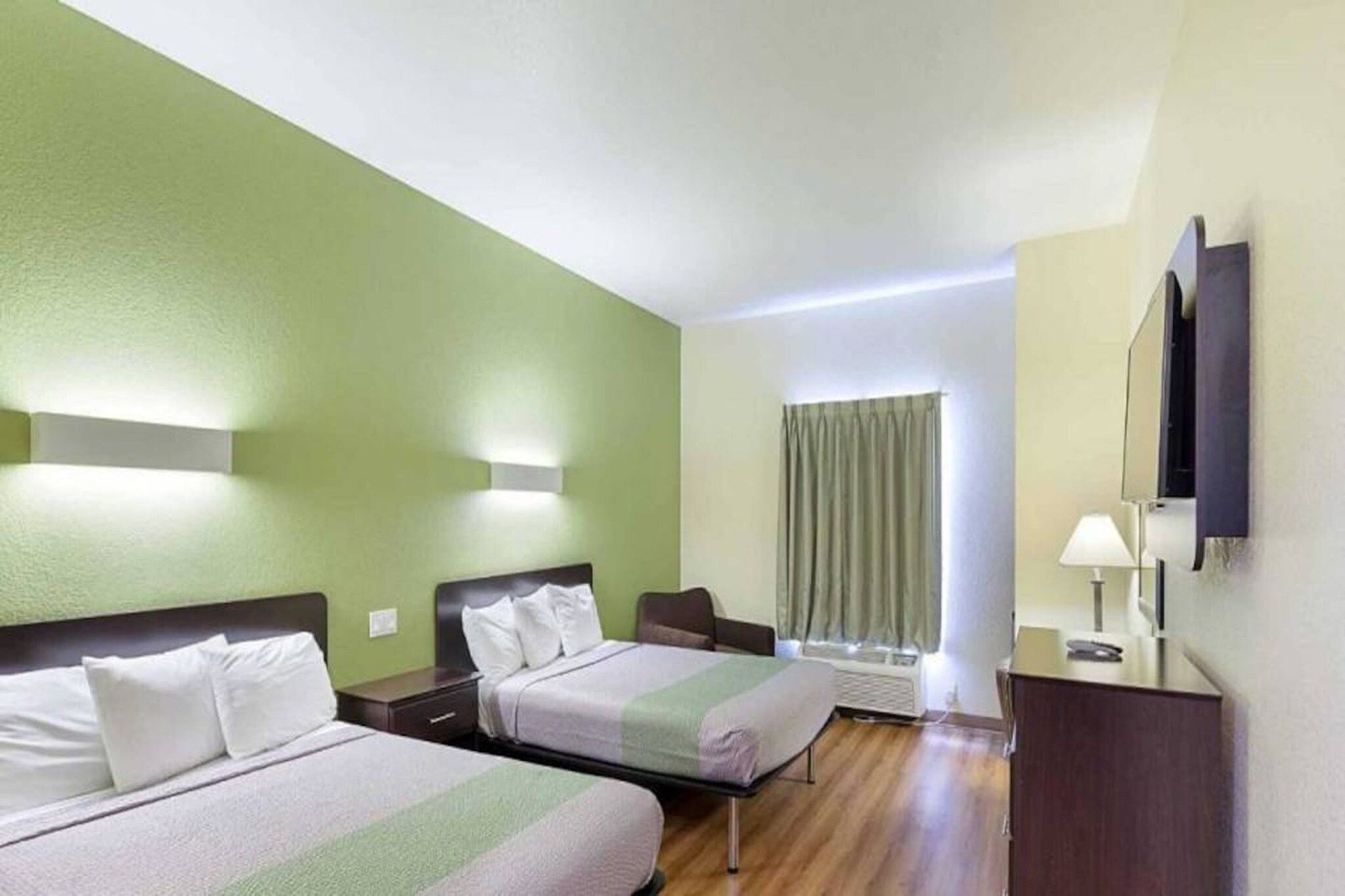 A hotel room with two beds, a desk, a chair, a window with curtains, and a flat-screen TV mounted on the wall.