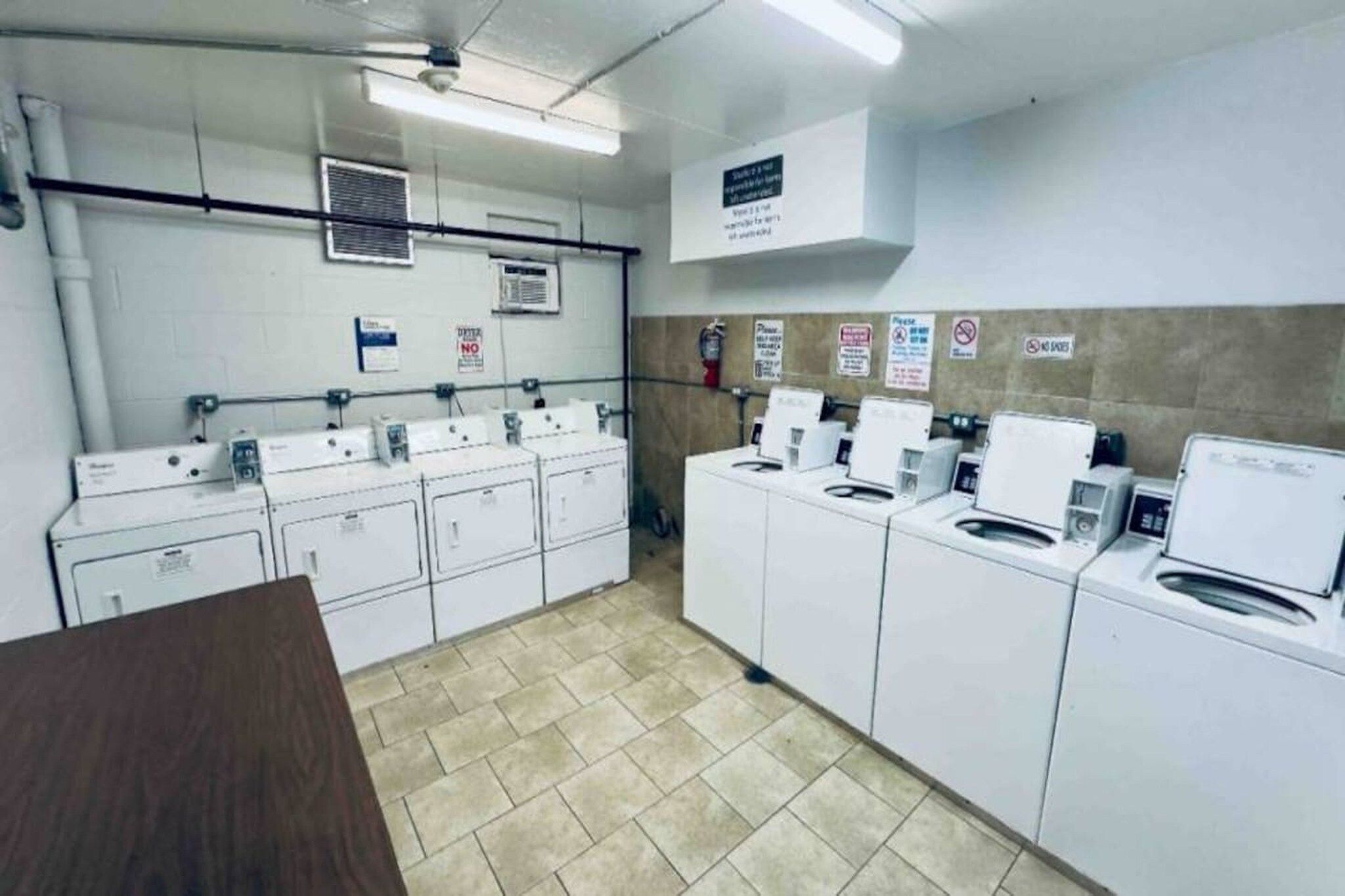 A laundry facility with multiple washing machines and dryers, a fire extinguisher, and safety signs on the walls.