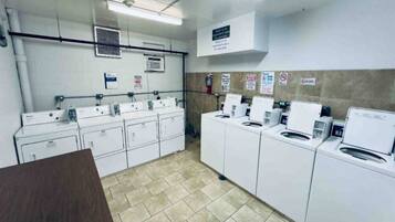 A laundry facility with multiple washing machines and dryers, a fire extinguisher, and safety signs on the walls.