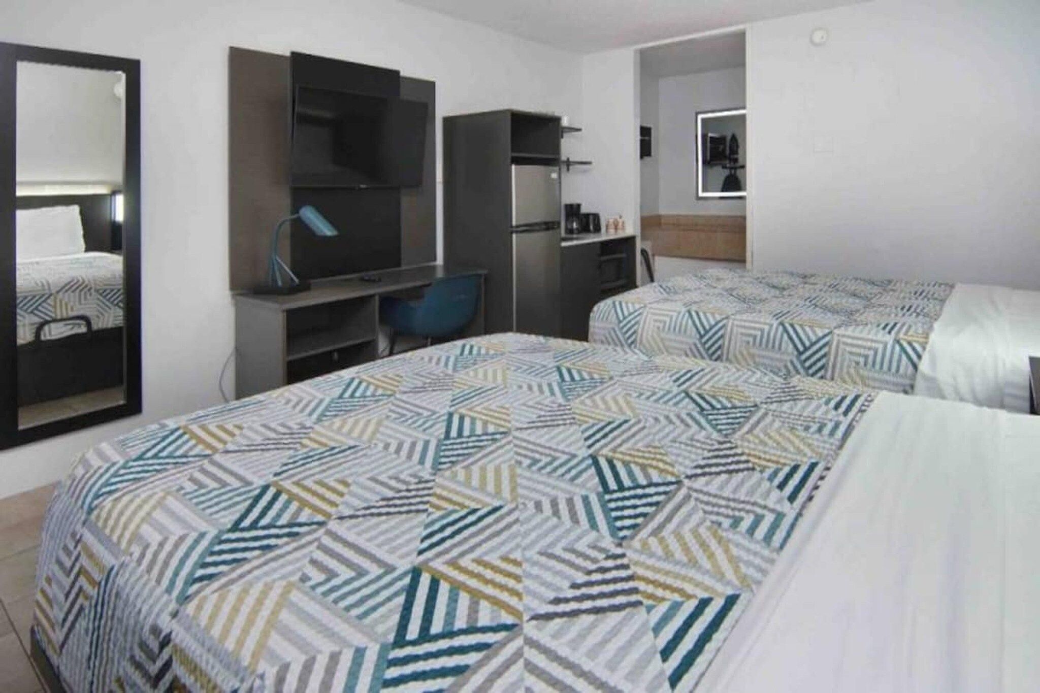 A hotel room with two beds, a desk, a chair, a mirror, and a kitchenette.