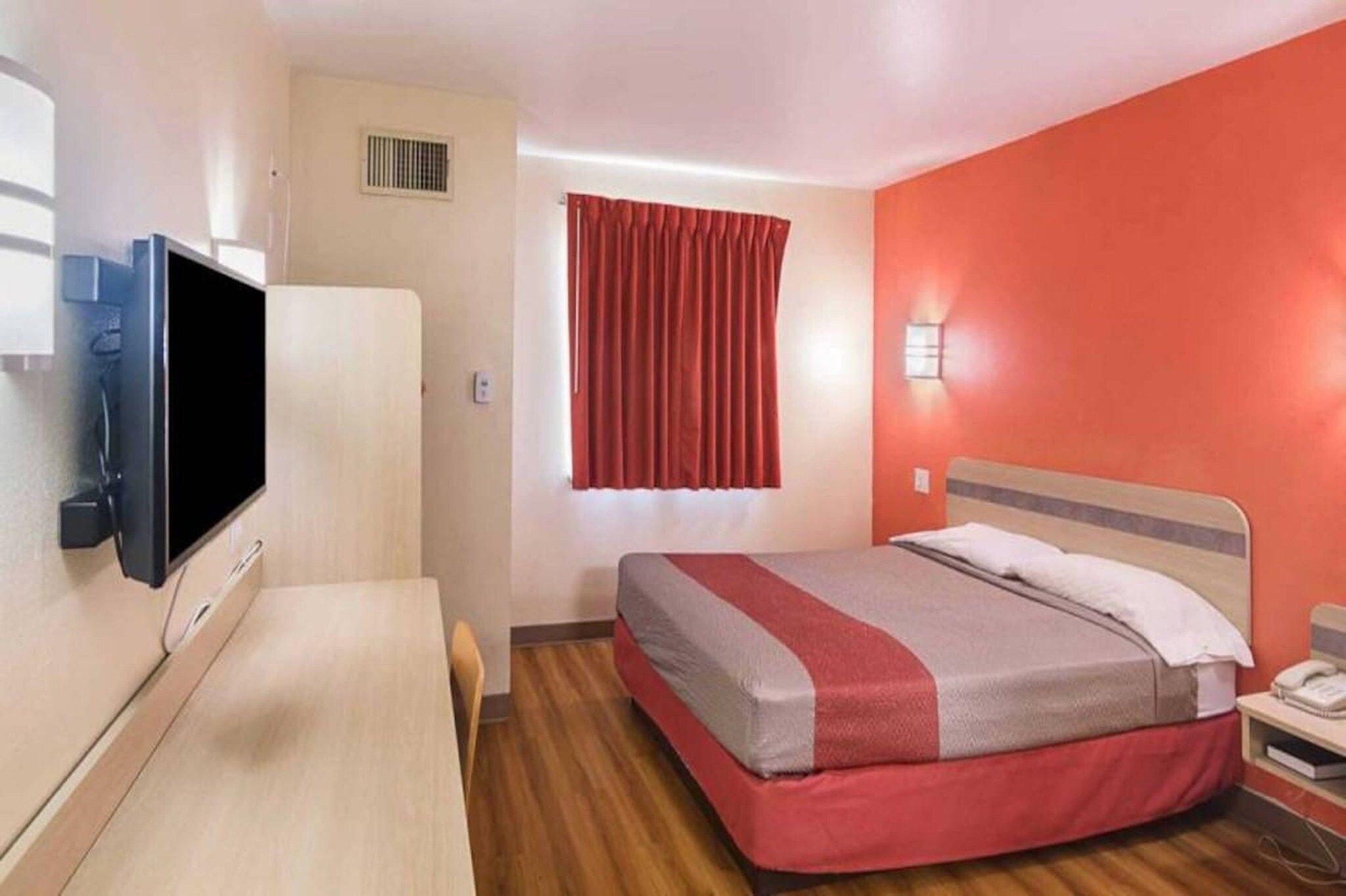 A hotel room with a bed, a desk, a television, a window with curtains, and a wall-mounted light.