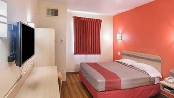 A hotel room with a bed, a desk, a television, a window with curtains, and a wall-mounted light.