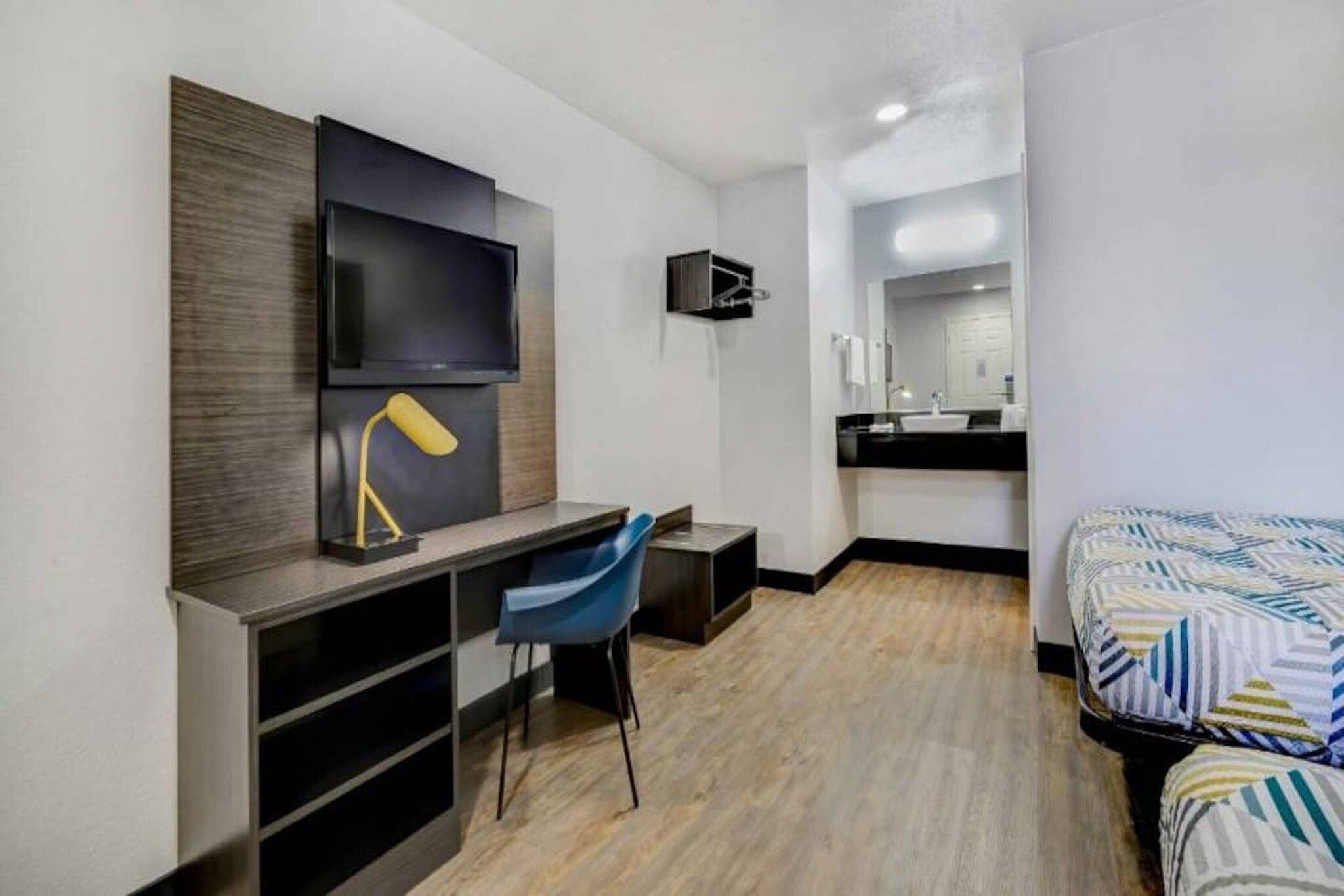 A modern hotel room with a flat-screen TV, a desk with a chair, a bed with patterned bedding, a bathroom with a sink and mirror, and a wooden floor.