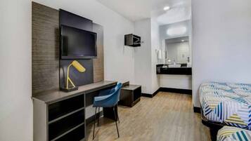 A modern hotel room with a flat-screen TV, a desk with a chair, a bed with patterned bedding, a bathroom with a sink and mirror, and a wooden floor.