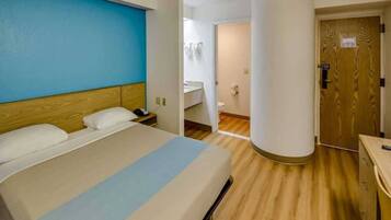 A hotel room with a bed, a bedside table, a nightstand, a wooden floor, a bathroom with a toilet and sink, and a door.
