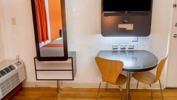 A room with a bed, a flat-screen TV mounted on the wall, a small table with two chairs, and a heater on the floor.