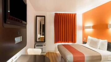 A hotel room with a bed, a desk, a chair, a television, a mirror, and a window with curtains.