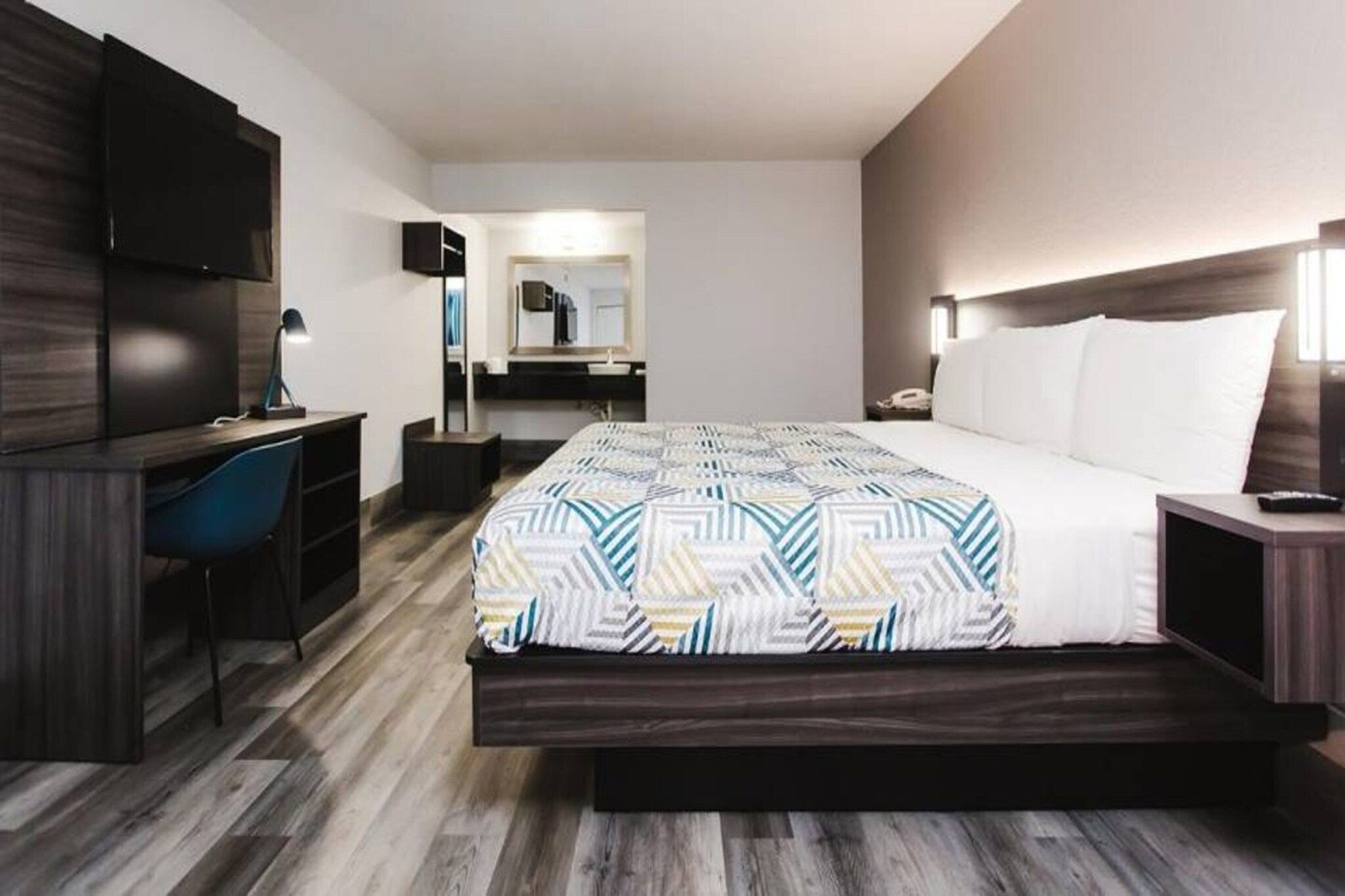 A hotel room with a bed, a desk, a chair, a TV, and a bathroom in the background.