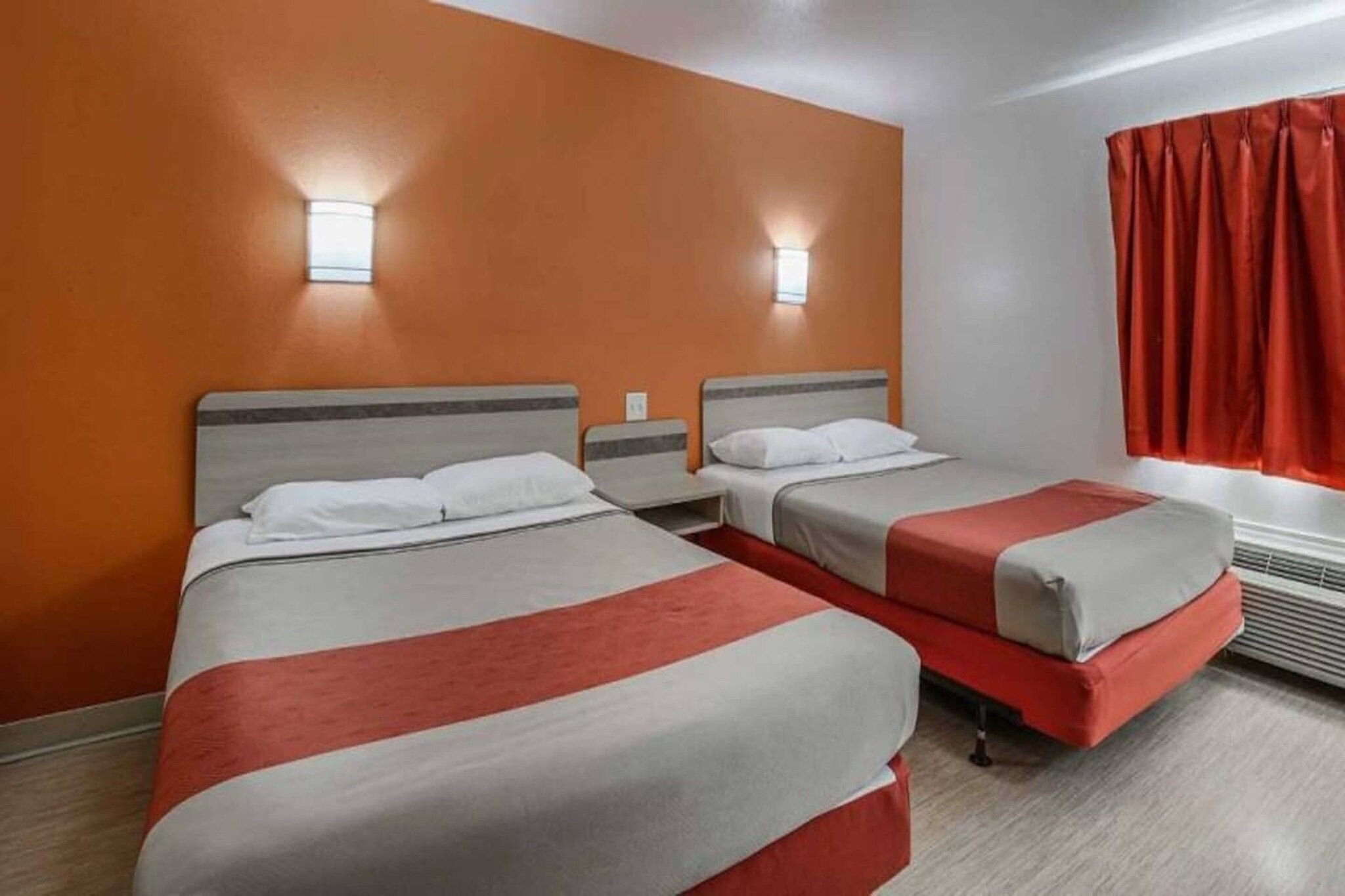 A hotel room with two beds, a window with red curtains, and a wall-mounted light fixture.