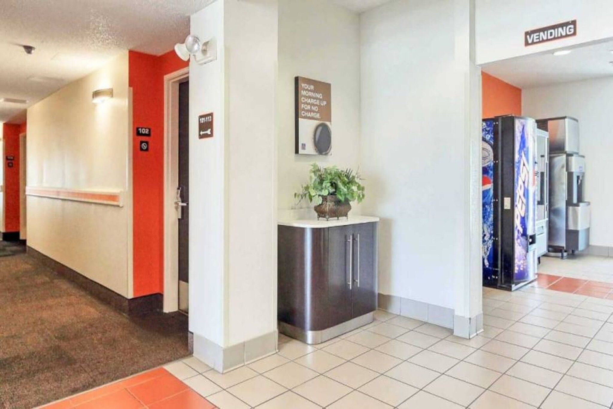 A hotel corridor with a vending machine, a plant on a counter, and a sign indicating a no-charge morning charge.