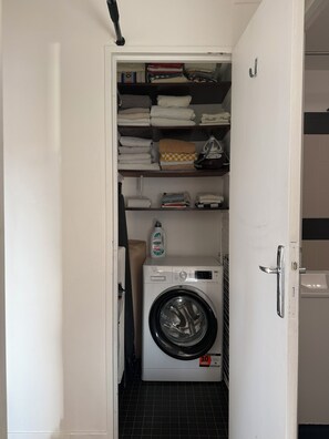 A laundry room with a front-loading washing machine, a shelf with folded towels, and a bottle of detergent. - Chic 75 m² Apartment with Views of the Canal and the Eiffel Tower (Paris)