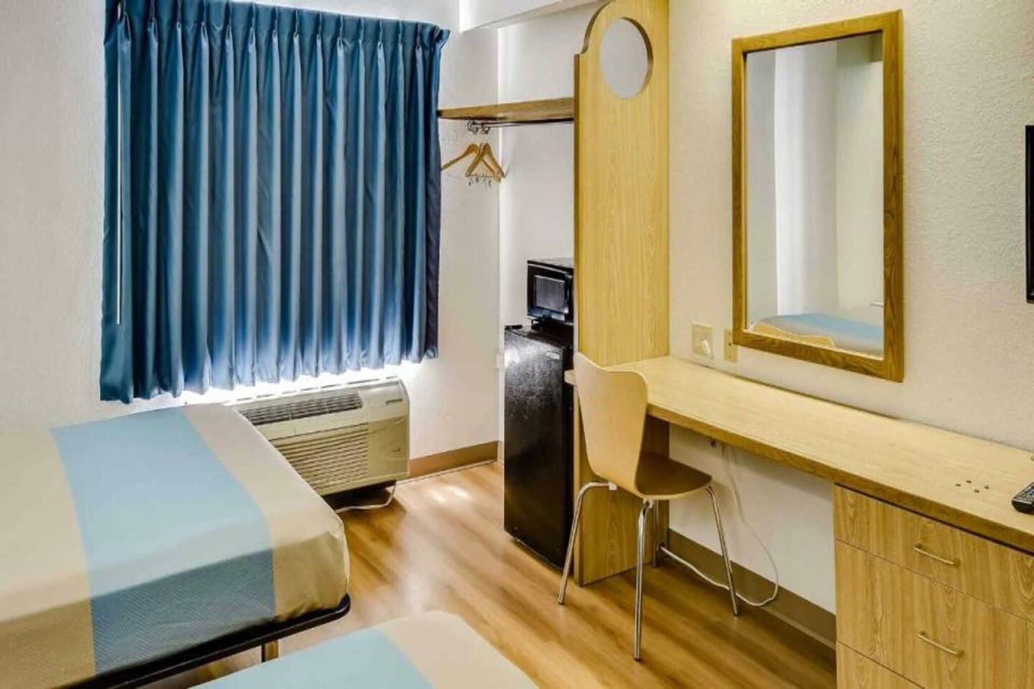 Une chambre d’hôtel comprenant un lit, un bureau, une chaise, un miroir, une armoire, une télévision et une fenêtre avec des rideaux.