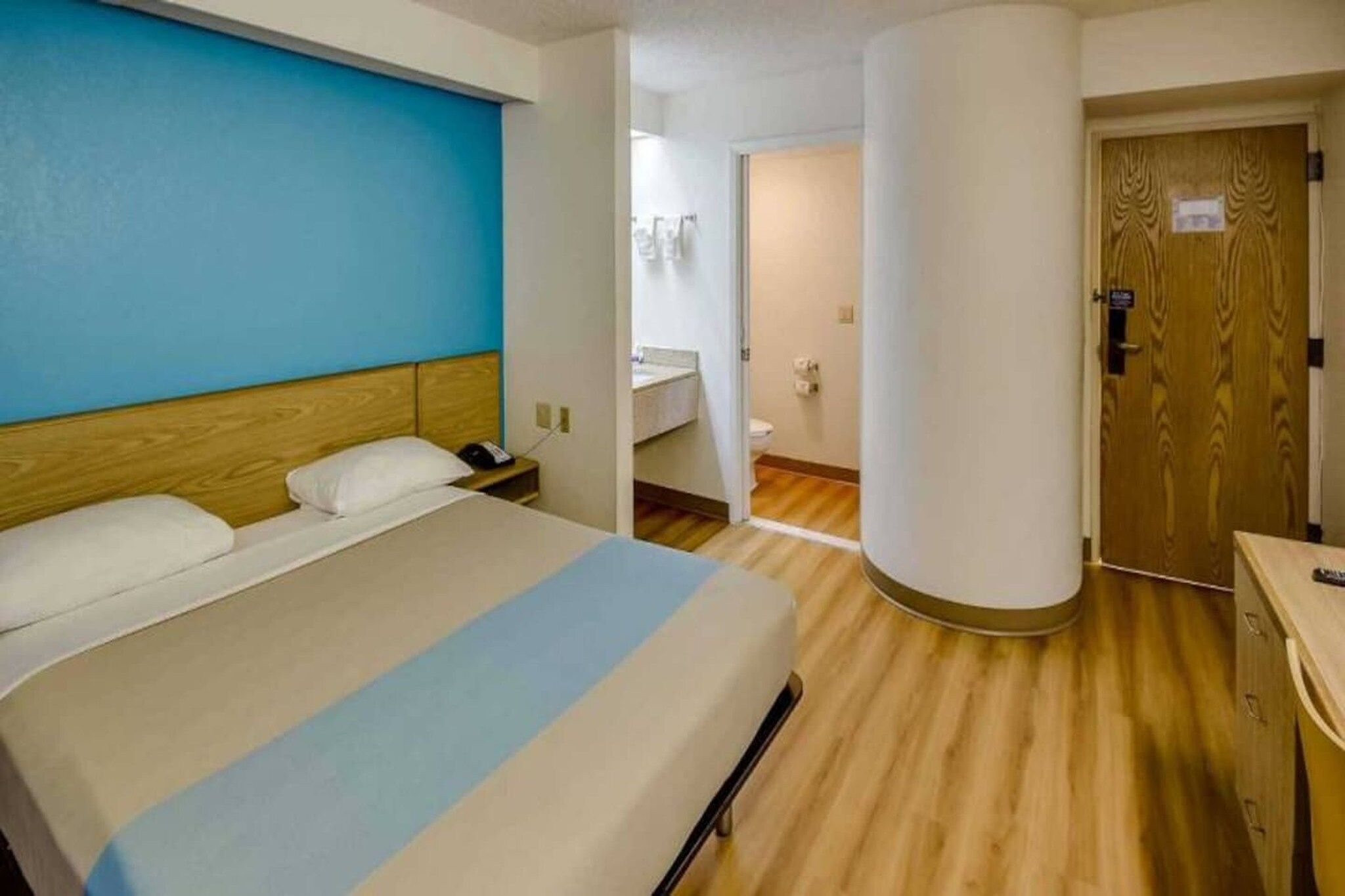 A hotel room with a bed, a bedside table, a nightstand, a wooden floor, a bathroom with a toilet and sink, and a door.