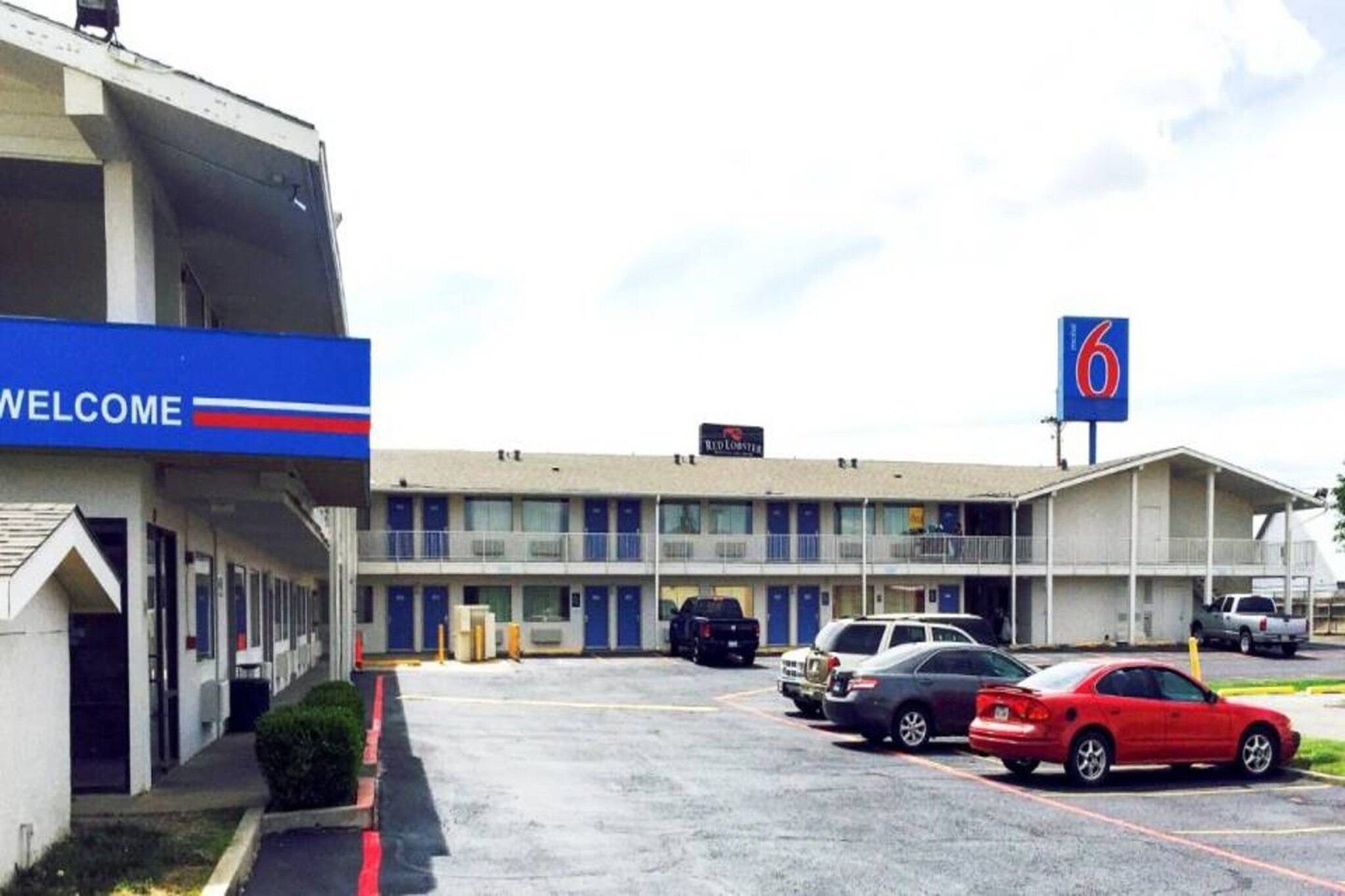 A motel with a 'WELCOME' sign, a '6' sign, and multiple cars parked in the lot.