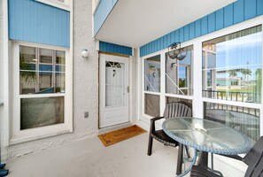 A covered porch with a glass table and chairs, a white door, and a blue exterior wall. - Walk to Beach & Heated Pool - Madeira Beach 2BR (Madeira Beach)