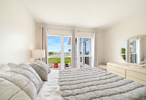 A bedroom with a large bed, a bedside table, a mirror, and a view of a green landscape through a sliding glass door.