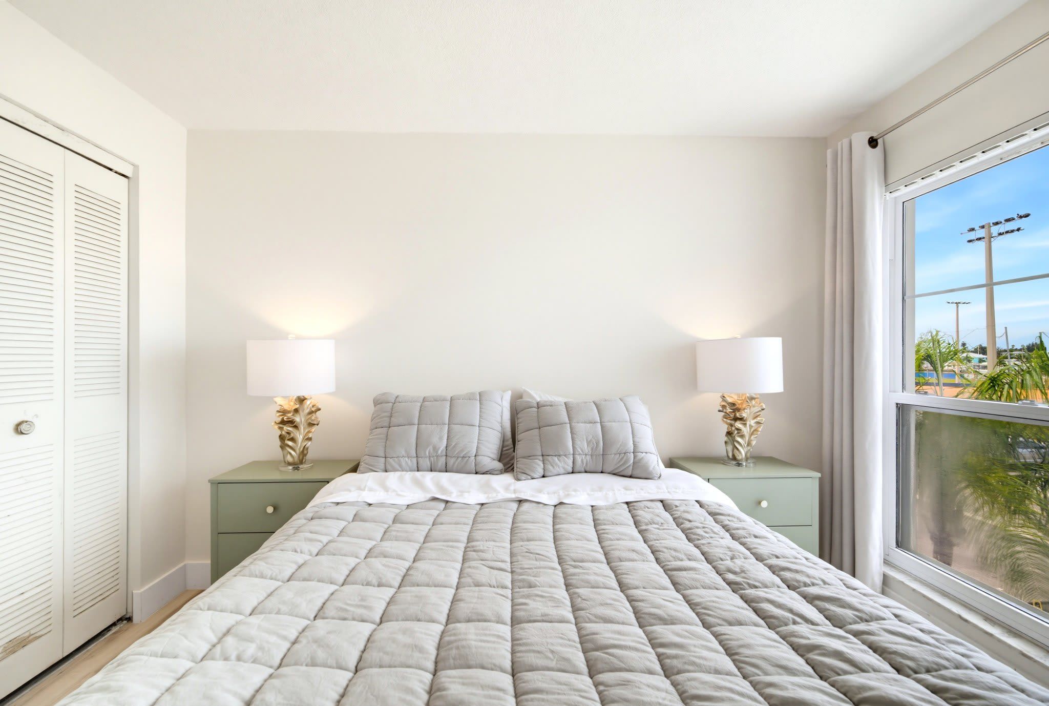 Un dormitorio con cama, dos mesitas de noche con lámparas, una ventana con vistas y una puerta con contraventanas.