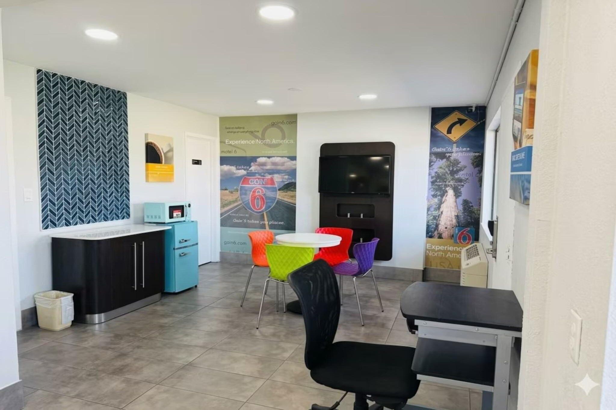 Een hotel receptie met een balie, een kleine kitchenette, kleurrijke stoelen en een groot poster aan de muur.