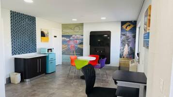 Een hotel receptie met een balie, een kleine kitchenette, kleurrijke stoelen en een groot poster aan de muur.