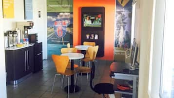 A cafe with a counter, chairs, and tables. There are posters on the wall and a computer on a stand.