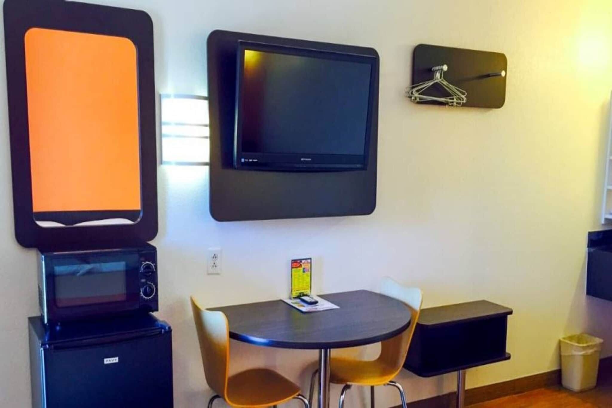 A room with a flat-screen TV mounted on the wall, a small table with two chairs, a microwave, and a trash bin.