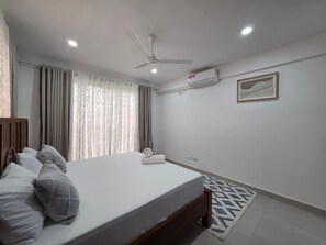 Une chambre à coucher avec un lit, un ventilateur de plafond, une fenêtre avec des rideaux et un tableau encadré au mur.