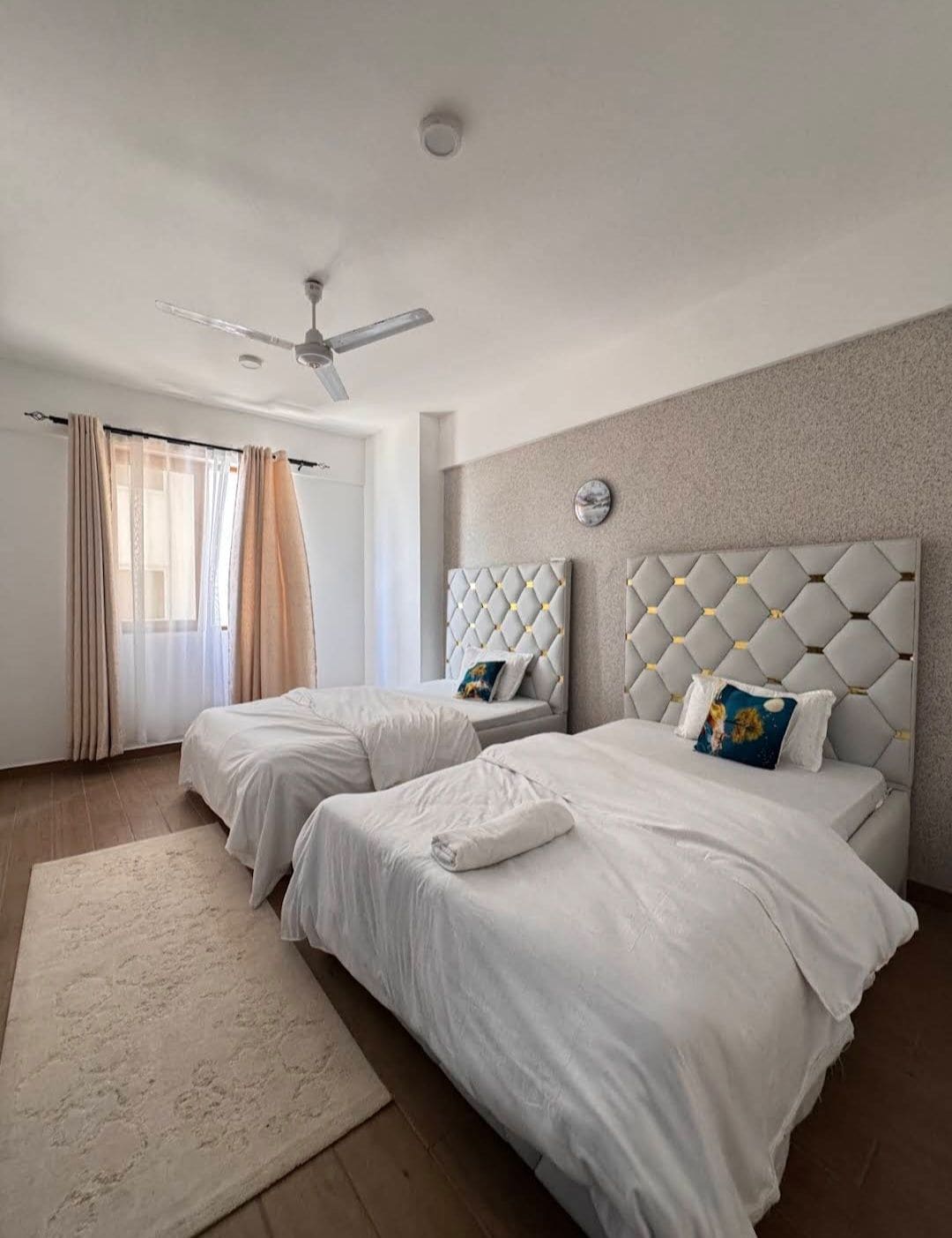 A hotel room with two beds, a ceiling fan, a large window with curtains, and a marble wall clock.