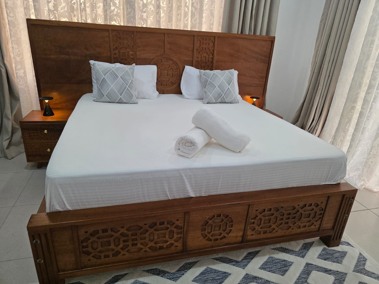 A neatly made bed with a wooden headboard, two bedside tables with lamps, and a patterned rug on the floor.