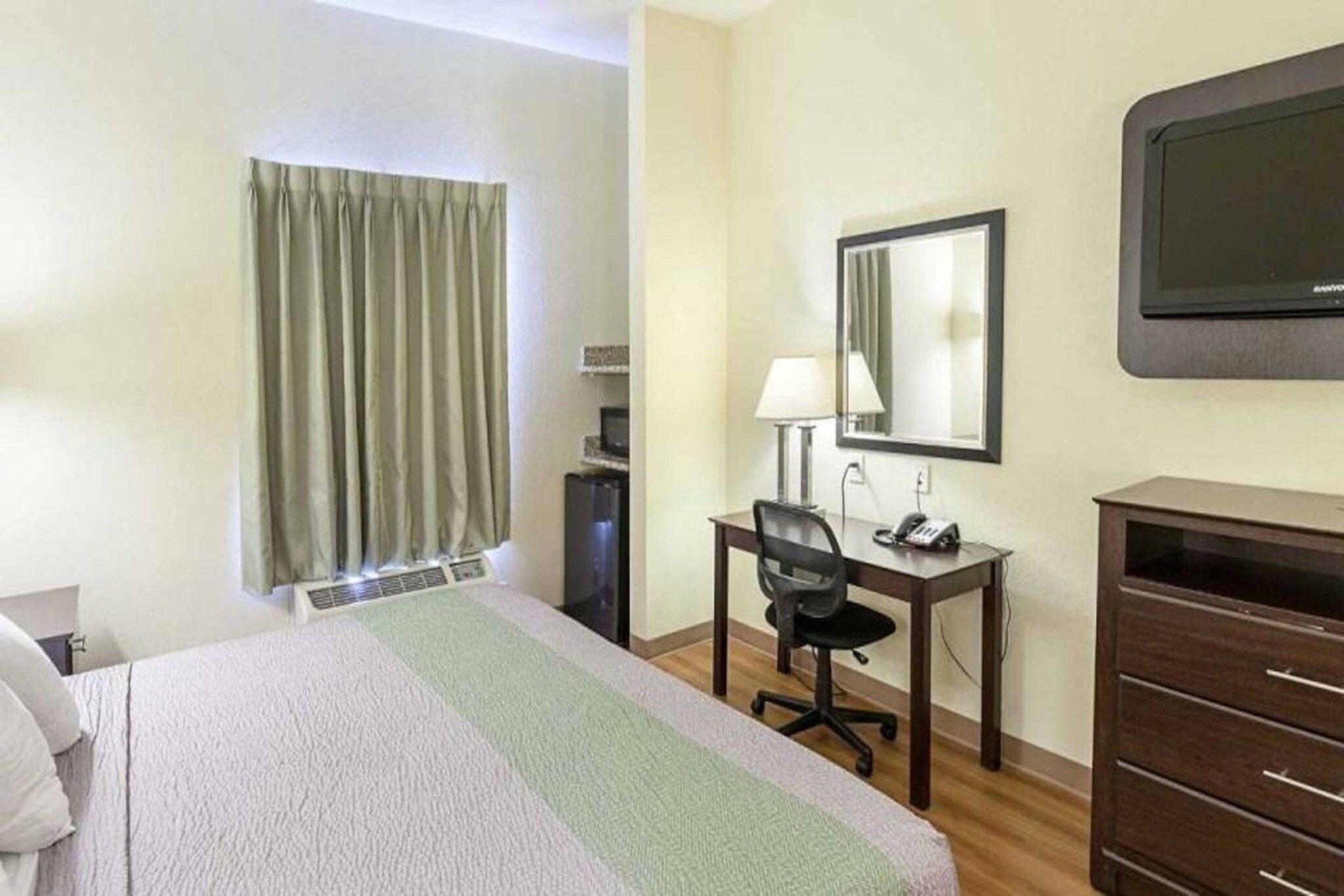 A hotel room with a bed, a desk with a chair, a dresser, a mirror, and a television on the wall.