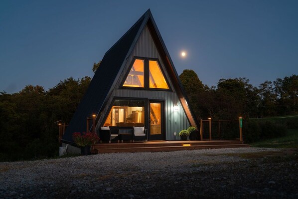 A modern A-frame house with a large glass window, a porch with seating, and a gravel driveway.