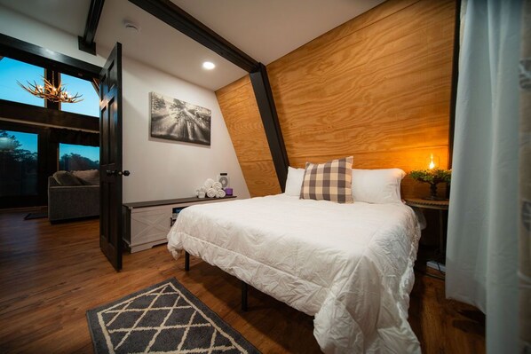 A bedroom with a large bed, a wooden headboard, a nightstand with a lamp, a painting on the wall, and a view of the outdoors through a door.