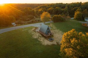 A single-story cabin with a porch, surrounded by a grassy area and trees, with a road in the background.