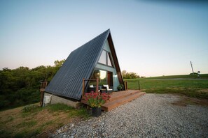 A modern, triangular-shaped building with a large glass window, a wooden deck, and a gravel driveway.