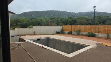 Una piscina con zona de grava alrededor, cerca, una valla de madera y vista a una colina boscosa.