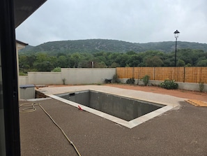 Una piscina amb una zona de grava al voltant, una tanca de fusta i vista a un turó forestal.