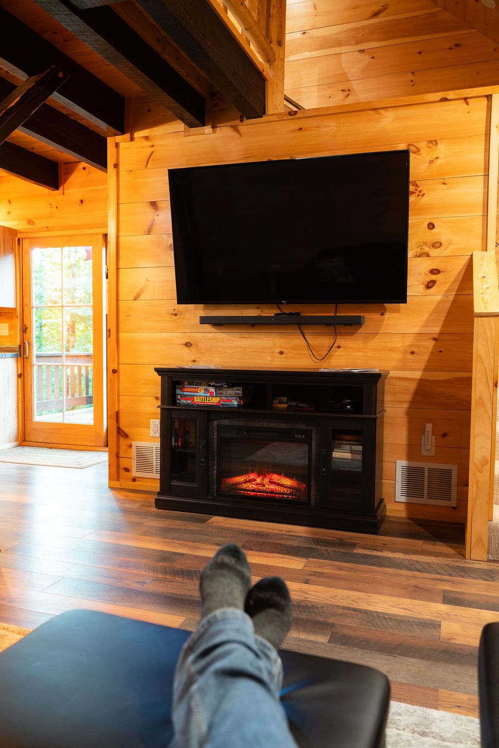 A wooden interior with a television mounted on the wall, a fireplace below, and a person relaxing on a couch.