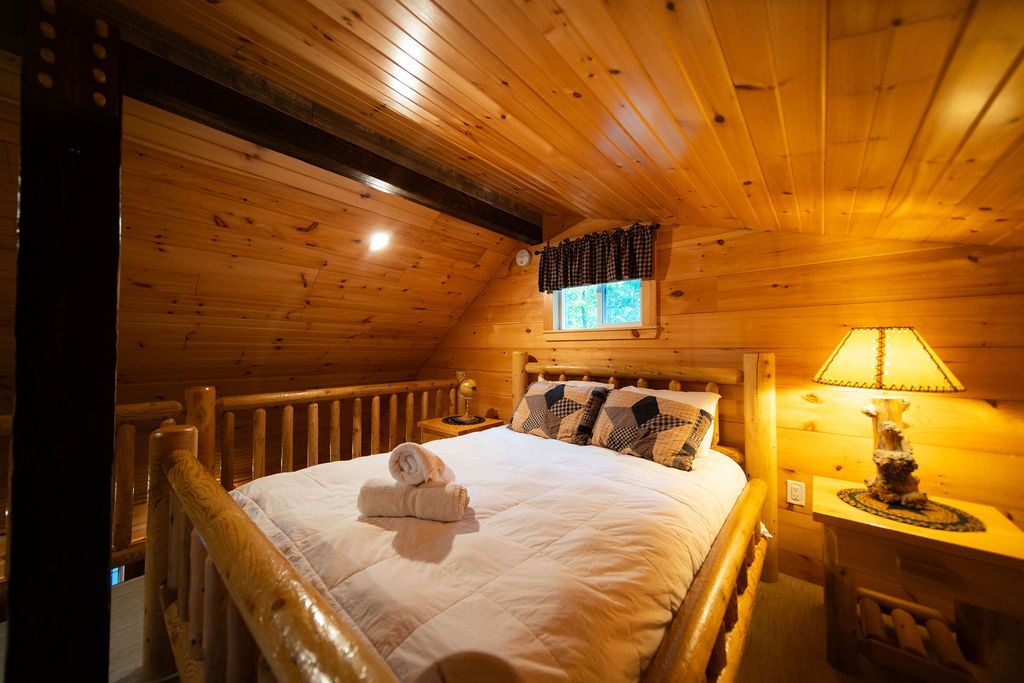 Ein Schlafzimmer einer Holzhütte mit einem Bett, einem Nachttisch, einer Lampe und einem Fenster mit Vorhängen.