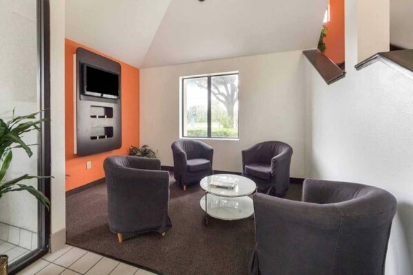 A modern living room with a flat-screen TV mounted on the wall, two armchairs, a round coffee table, and a large window providing a view of the outdoors.