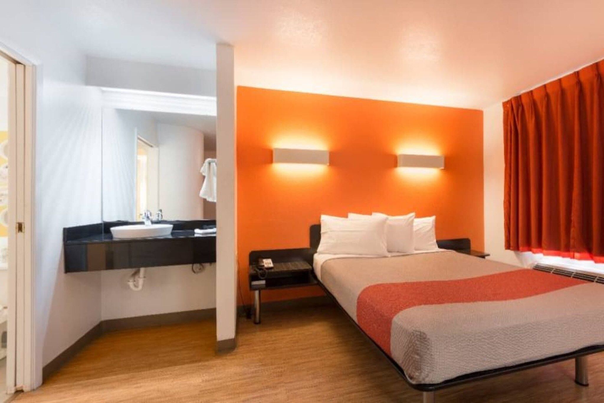 A hotel room with a bed, a nightstand, a bedside table, a bathroom with a sink and mirror, and orange walls.