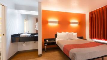 A hotel room with a bed, a nightstand, a bedside table, a bathroom with a sink and mirror, and orange walls.