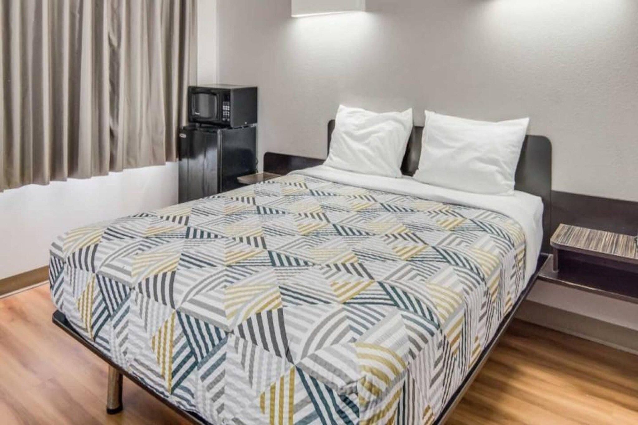 A hotel room with a bed, a nightstand, a microwave, and a window with curtains.