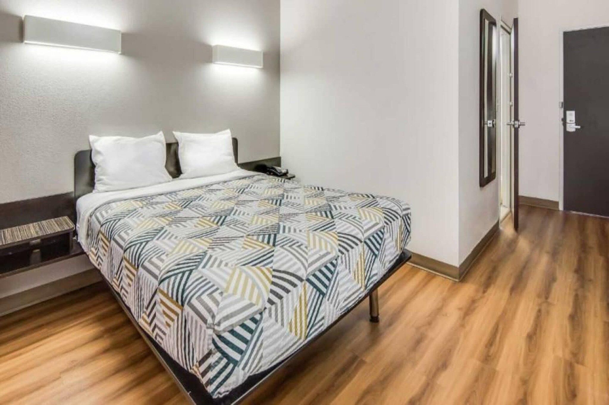 A hotel room with a bed, a nightstand, a wall-mounted light, and a door.