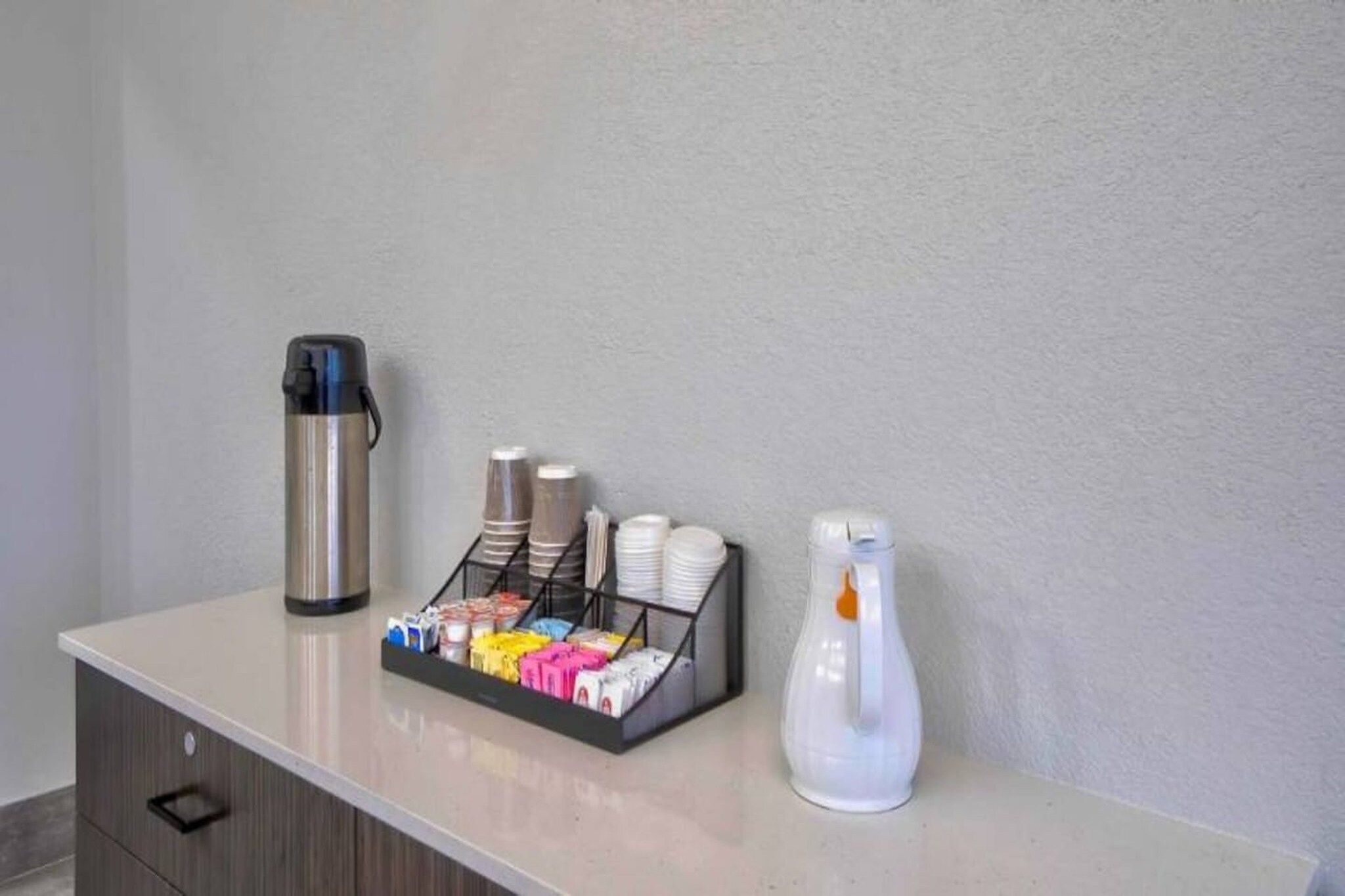Área de recepção de hotel com estação de café, incluindo máquina de café, dispensador de água e escorredor com xícaras e pratos.