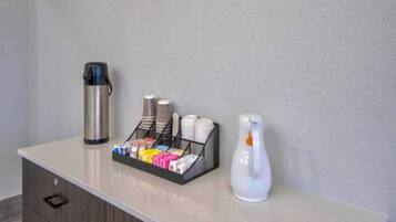 Área de recepção de hotel com estação de café, incluindo máquina de café, dispensador de água e escorredor com xícaras e pratos.