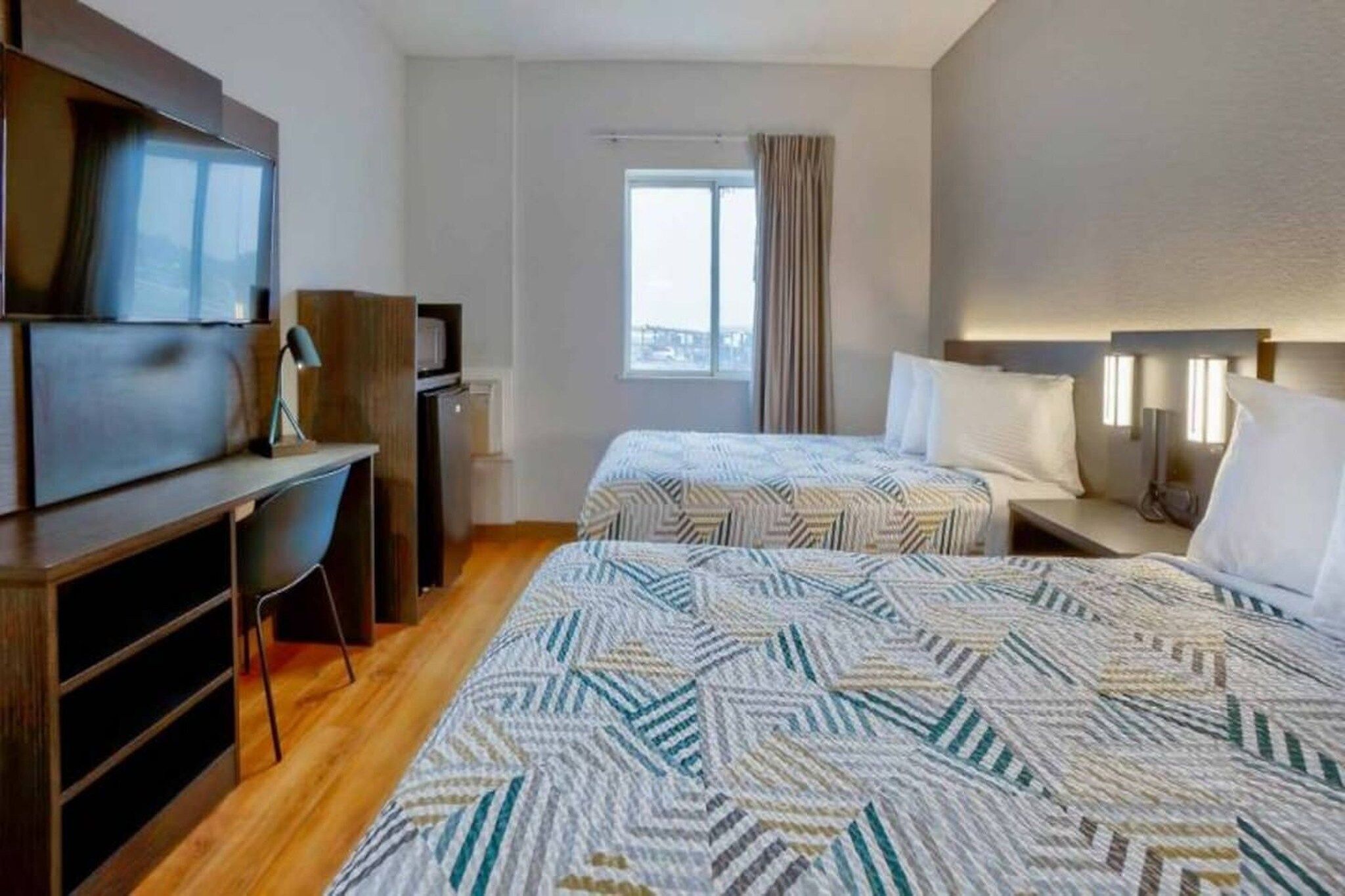 A hotel room with two beds, a desk, a television, and a window with curtains.