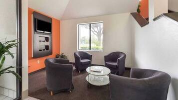 A modern living room with a flat-screen TV mounted on the wall, two armchairs, a round coffee table, and a large window providing a view of the outdoors.
